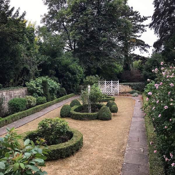 Formal English garden with clipped box shapes, a lattice gate, and flowering shrubs along a central walkway.