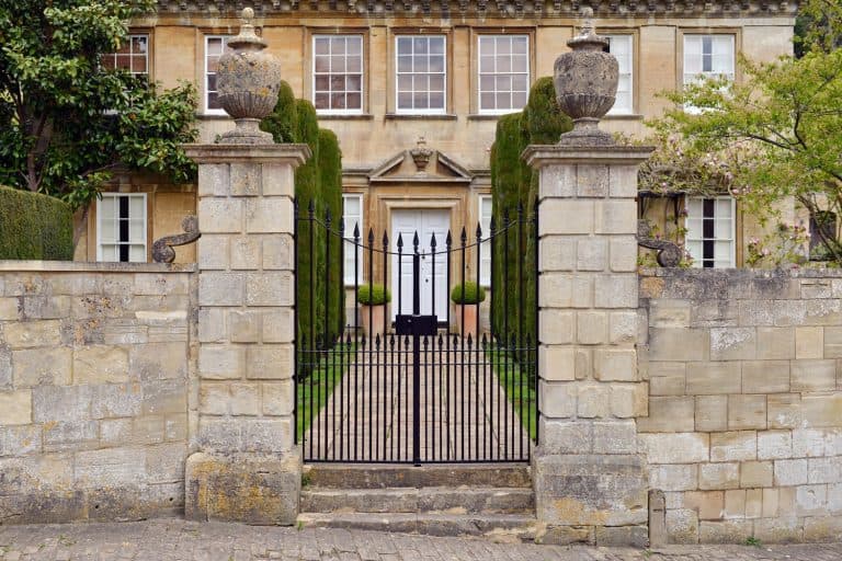 Ornate black iron gate with sharp spikes and tall stone pillars, flanked by symmetrical potted plants, leading to a well-maintained property.
