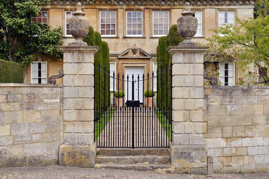 Ornate black iron gate with sharp spikes and tall stone pillars, flanked by symmetrical potted plants, leading to a well-maintained property.