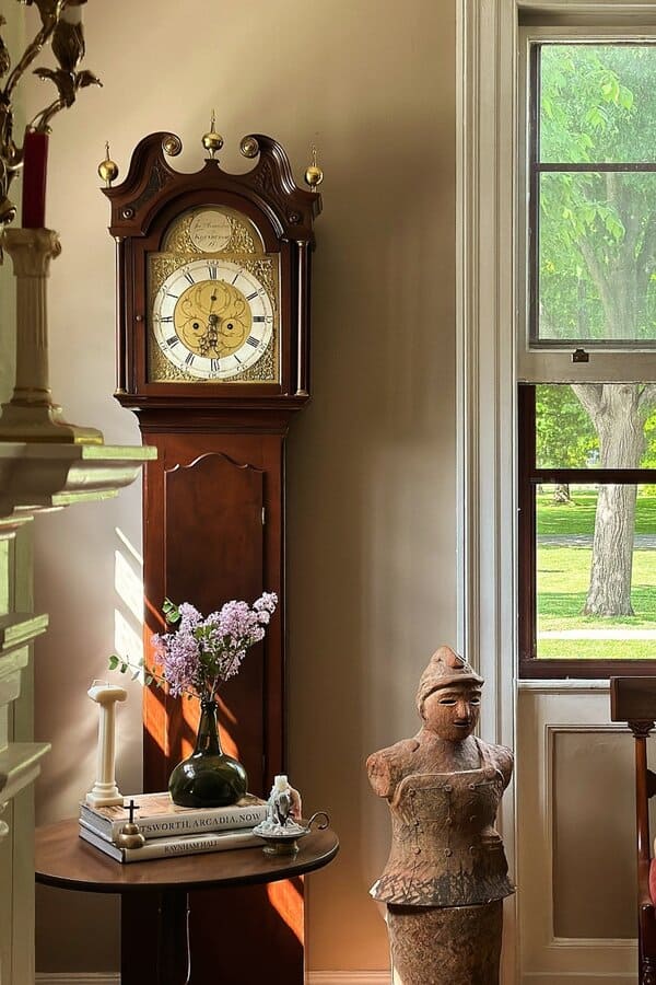 A grandfather clock, a seated dog looking out of a window, a statue, and a vase with flowers on a small table.