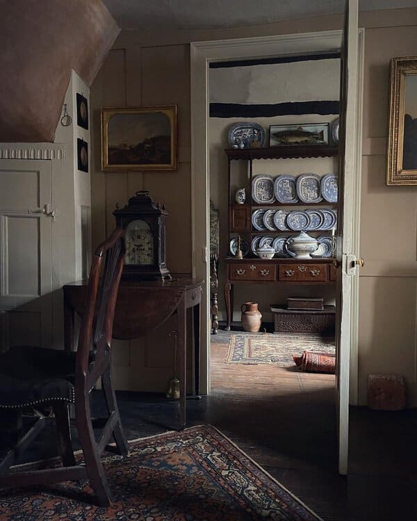 Dimly lit interior with paneled walls, an antique clock, and a dresser filled with blue-and-white plates for a cozy, historic feel.