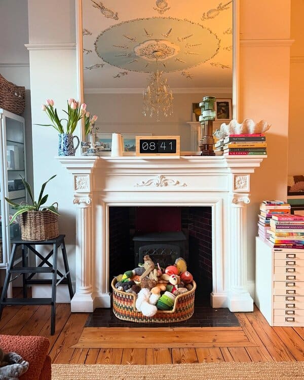 White Georgian fireplace topped by a tall mirror, with a basket of children’s toys in the hearth and warm wooden floors underfoot.