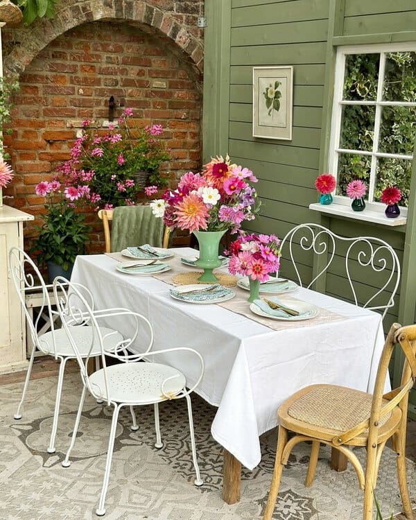 Outdoor dining area with colorful flowers, white metal chairs, and a brick arch backdrop for a cheerful garden setting.