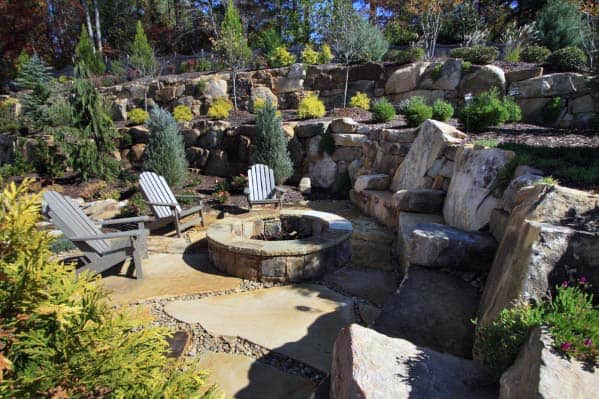 Stone fire pit with Adirondack chairs surrounded by rocks and greenery in a terraced garden setting