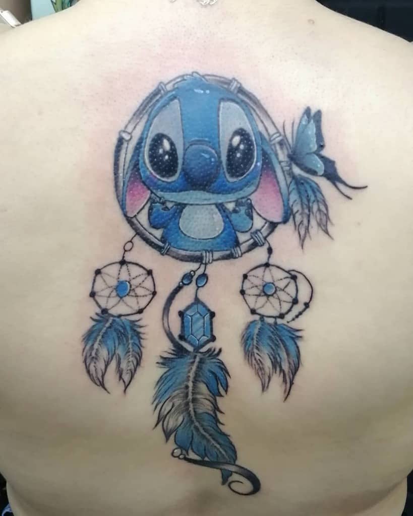 Colorful tattoo of Stitch from Lilo & Stitch inside a dreamcatcher, surrounded by blue feathers, beads, and a butterfly, on the upper back.