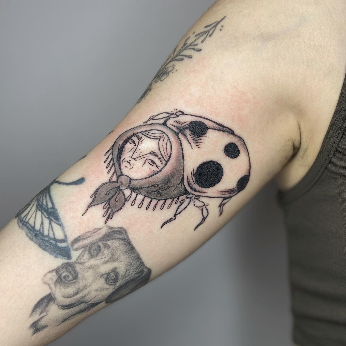 Black and grey tattoo of a ladybug with a human-like face in a headscarf, blending surrealism and traditional tattoo style.