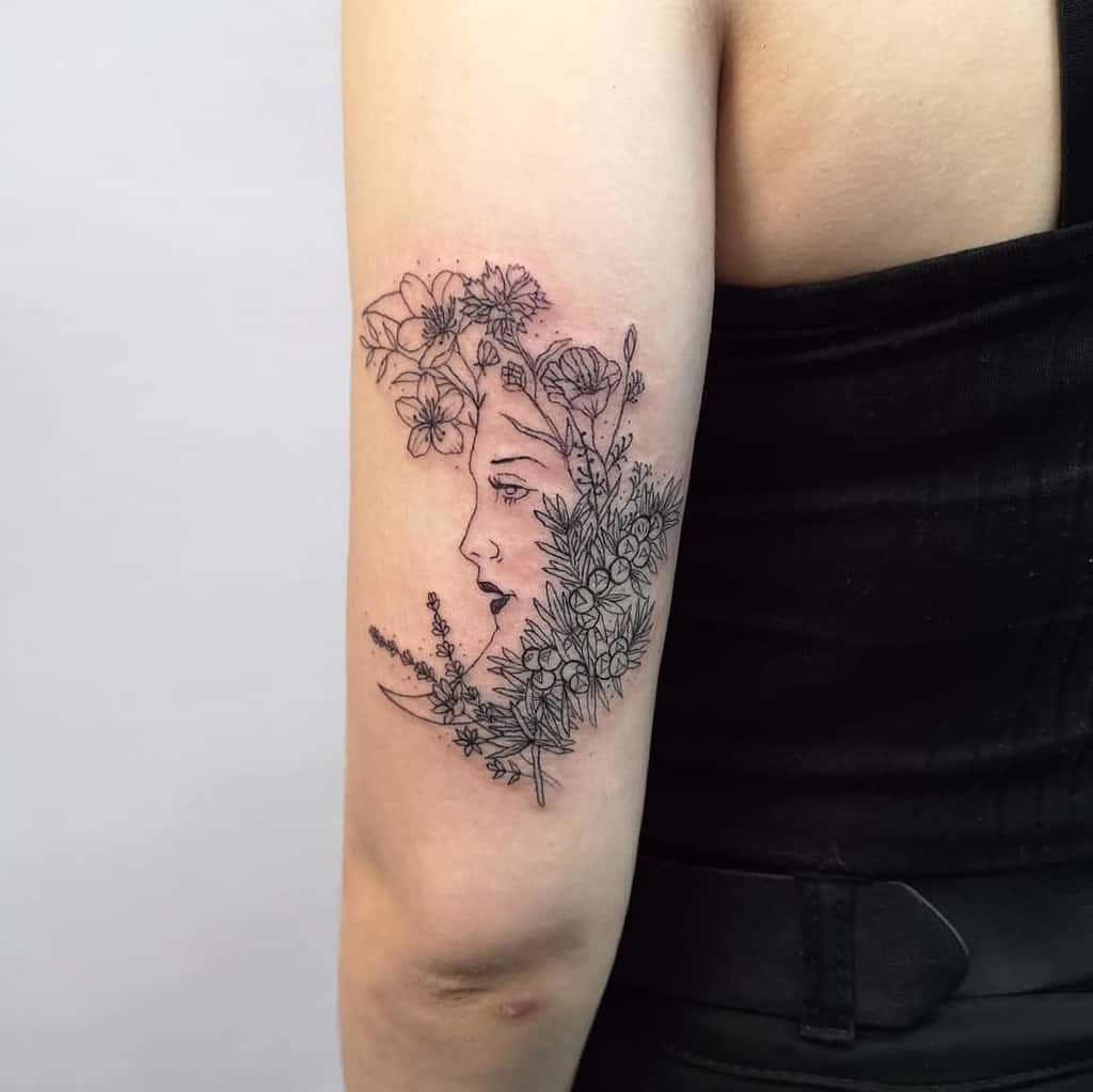 A tattoo on the upper arm featuring the profile of a woman surrounded by a bouquet of flowers, including lavender and other floral elements, in fine black line art.