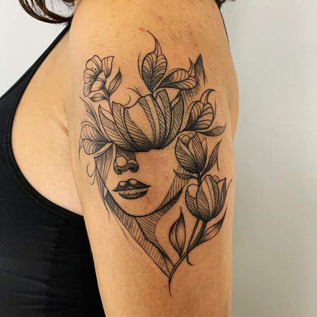 A surreal black-line tattoo on the upper arm, featuring a woman's face partially obscured by blooming tulips and intricate floral details.