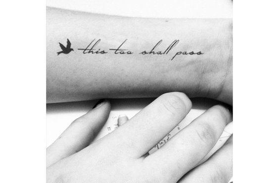 A "This Too Shall Pass" tattoo in cursive, with a bird design, on a person's forearm.