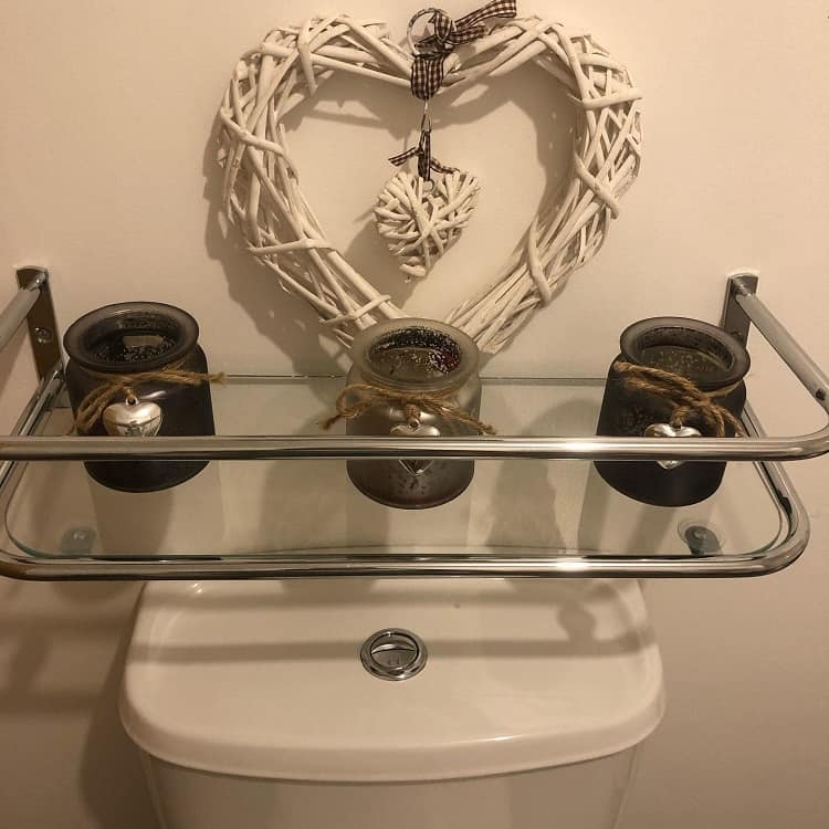Glass and metal shelf over the toilet with decorative candles and heart-shaped wicker ornament.