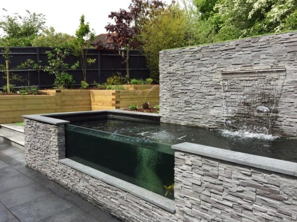 Rustic stone waterfall feature with a glass-paneled pond in a modern landscaped backyard.