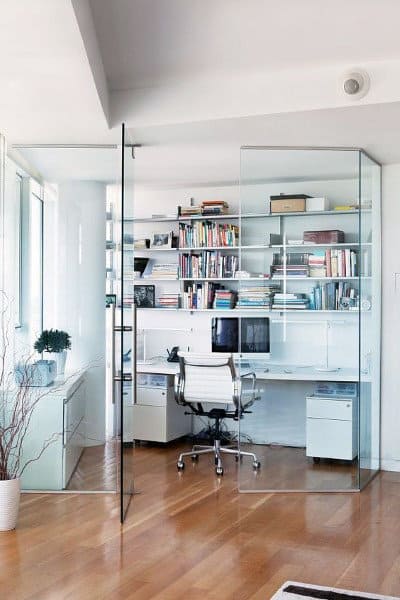A modern home office with glass walls, wooden flooring, bookshelves, and a sleek white desk and chair