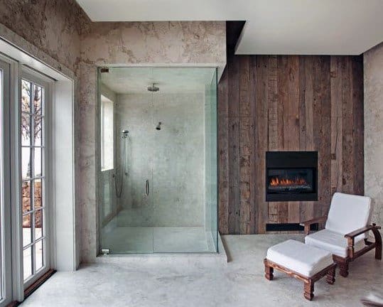 Rustic bathroom with a glass shower, fireplace, and a cushioned chair with ottoman