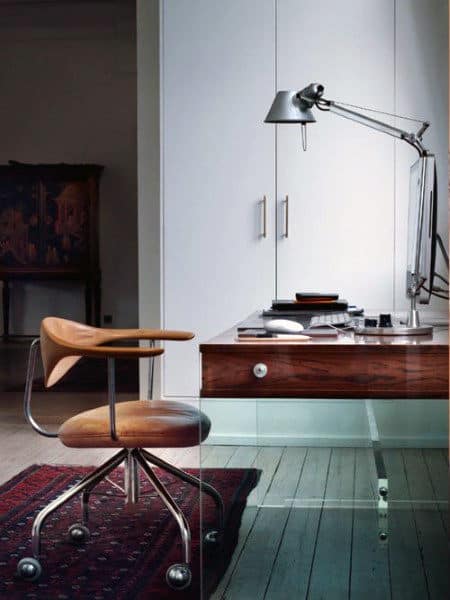 Modern home office with a wooden chair, glass desk, desk lamp, and shelves in the background