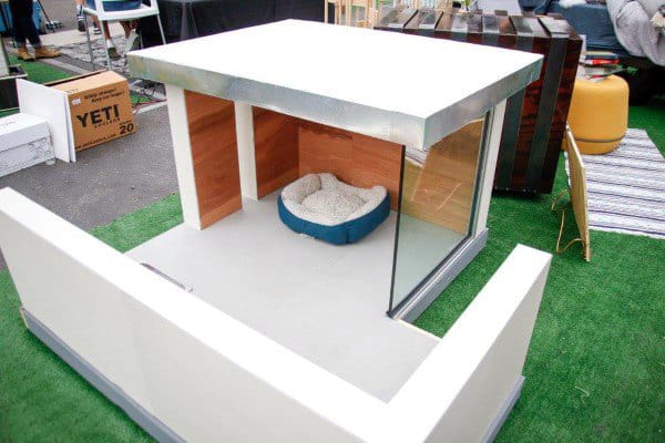 A modern dog house with glass walls and a sleek design, featuring a small dog bed, food bowl, and tennis ball outside