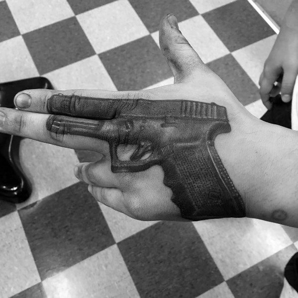 Realistic black and grey tattoo of a Glock pistol on the hand, positioned so that the index and middle fingers mimic the barrel of the gun.