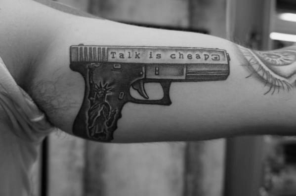 Black and grey tattoo of a Glock pistol on the inner arm, featuring "Talk is cheap" engraved on the slide and a Statue of Liberty design on the grip.