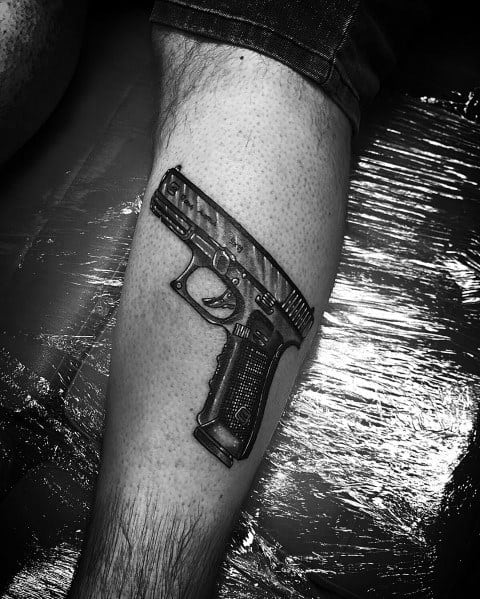 Realistic black and grey tattoo of a Glock pistol on the lower leg, featuring intricate shading and texture for a three-dimensional effect.