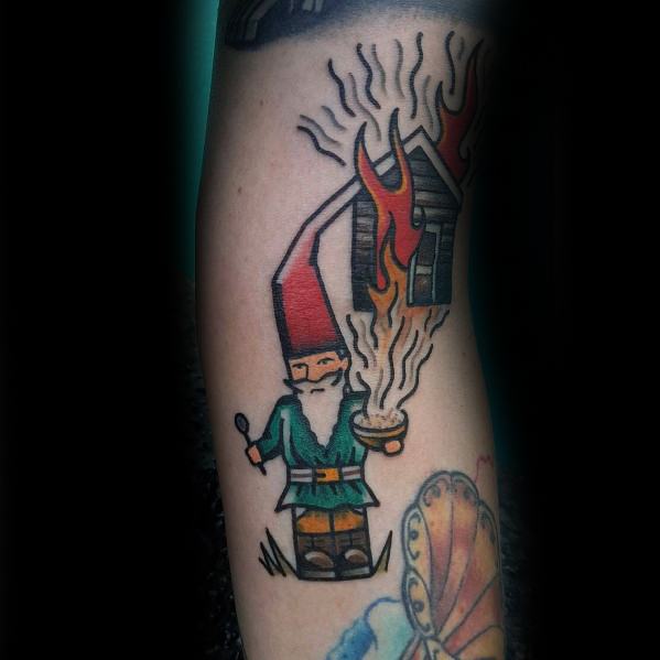 Tattoo of a gnome with a red hat and green outfit holding a smoking bowl, with a burning house in the background, done in a bold, cartoon style.