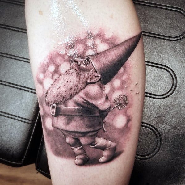 Black and white tattoo of a gnome with a long beard and pointed hat, holding a dandelion and blowing its seeds into a dreamy, blurred background.