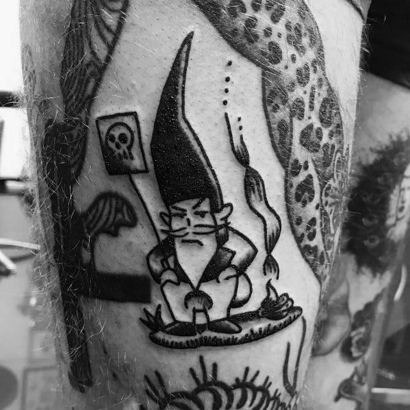 Black and white tattoo of a grumpy gnome with a tall hat, sitting on a mushroom, smoking a pipe, with a skull sign in the background.