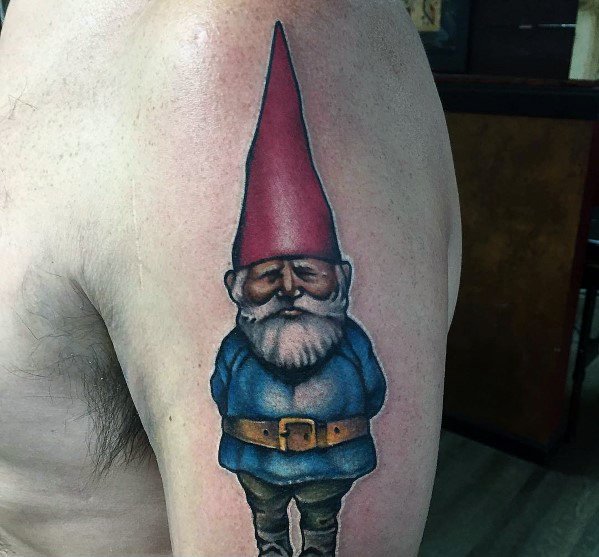Tattoo of a serious-looking gnome with a tall red hat, blue coat, and brown boots, standing with hands behind his back on the upper arm.