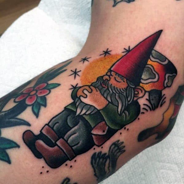 Tattoo of a peaceful gnome with a red hat and green outfit, lying down with hands clasped, resting against a mushroom with a sunset background.