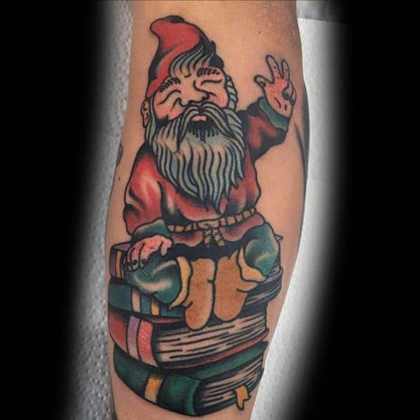 Tattoo of a wise gnome with a red hat and long beard, sitting cross-legged on a stack of books, flashing a peace sign with a playful smile.