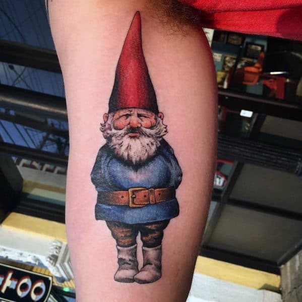 Tattoo of a classic garden gnome with a tall red hat, white beard, and blue coat, standing with hands behind his back and a slight smile.