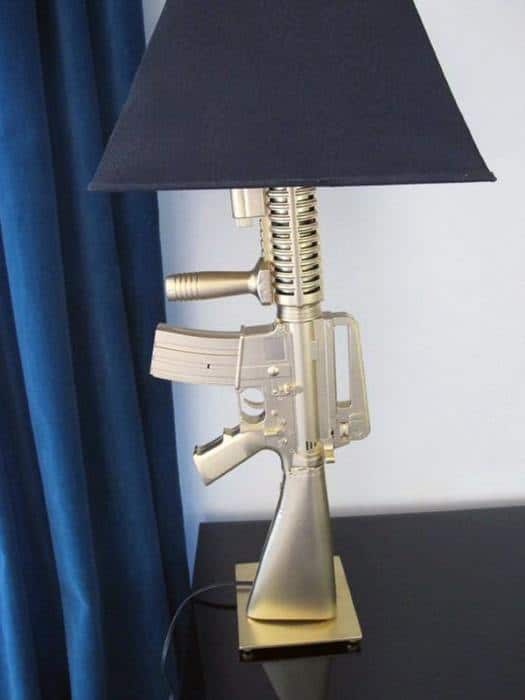 A lamp designed like a gold rifle with a black lampshade, placed on a dark table near blue curtains