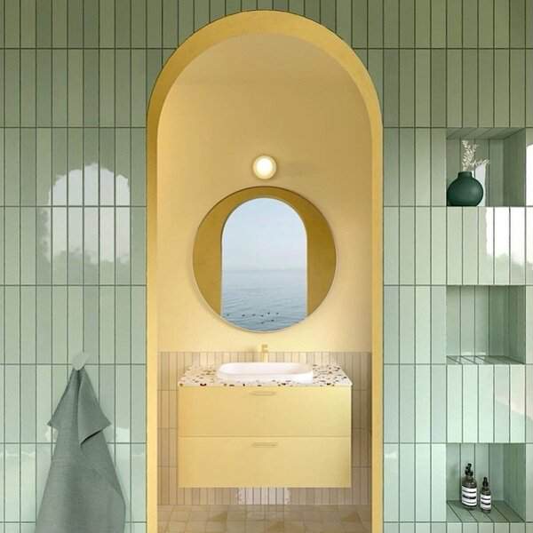 A yellow arch vanity with a round mirror reflecting ocean views, flanked by green tile and terrazzo counters.
