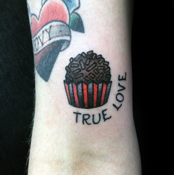 Tattoo of a chocolate truffle in a red-striped wrapper with "TRUE LOVE" written below, symbolizing a passion for sweets.