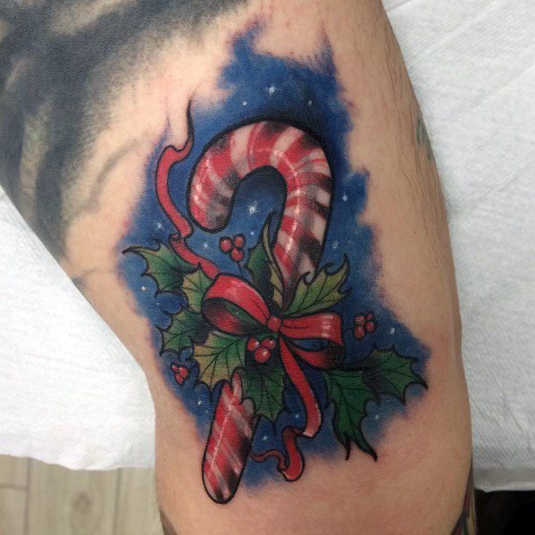 Christmas tattoo of a candy cane wrapped in a red ribbon, surrounded by holly leaves and berries, set against a starry blue background for a festive look.