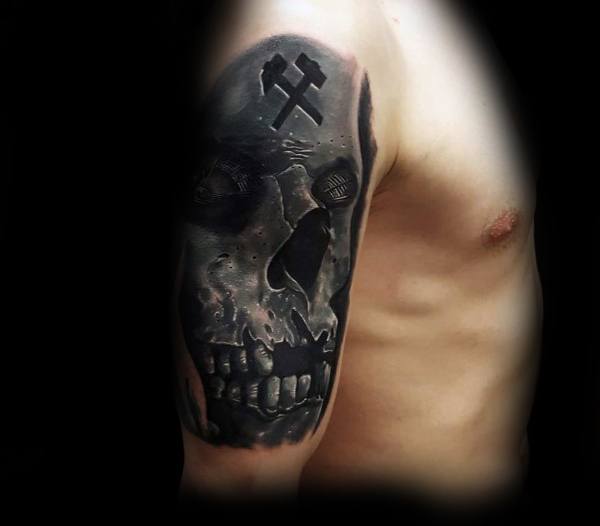 Dark and intense black and grey tattoo of a skull with a miner’s pickaxe symbol carved into the forehead on the upper arm.
