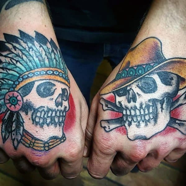 Tattoo of two skulls on each hand, one wearing a Native American headdress and the other a cowboy hat with crossed bones, done in bold traditional style.