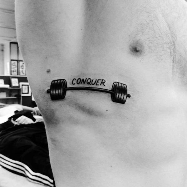 Minimalist barbell tattoo on the ribs with the word "CONQUER," symbolizing strength, perseverance, and dedication to fitness and self-improvement.