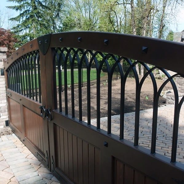 Dark brown wooden driveway gate with black arched metal bars and decorative rivets.