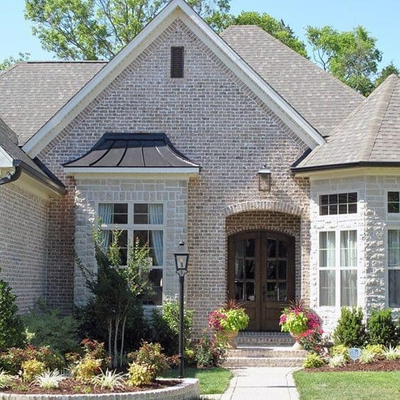 Charming home with light brick and stone exterior, arched wooden door, metal awning, and vibrant landscaping for a welcoming entry