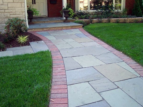 Walkway with a brick border leads to a house entrance, surrounded by grass and plants