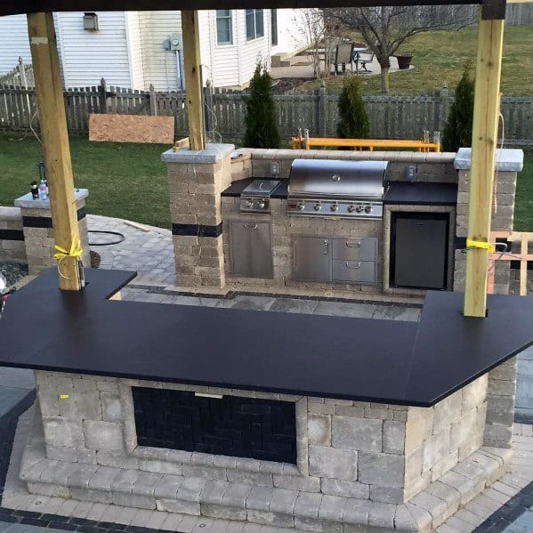Outdoor kitchen with stone countertops, built-in grill, and stainless steel appliances under a wooden pergola