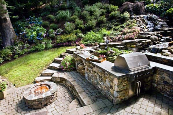 Stone patio with a built-in grill and fire pit, surrounded by lush greenery and a cascading waterfall