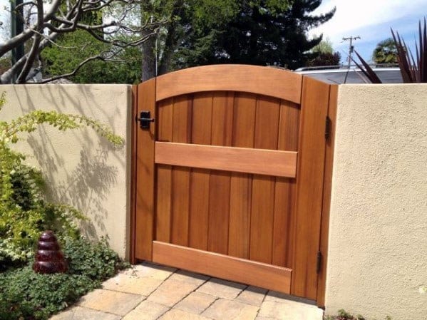 Wooden gate with an arched top set in a beige stone wall, surrounded by plants, on a paved path