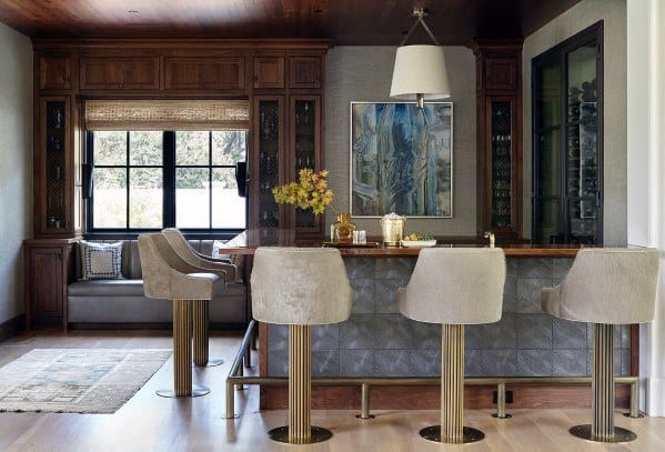 Luxurious wet bar with brass foot rail, tufted bar front, and elegant cream barstools on light wood flooring.