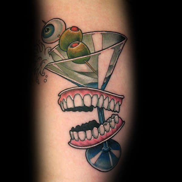 Surreal martini glass tattoo featuring olives, an eyeball, and a set of dentures biting the stem. A bold and quirky design with vivid details.