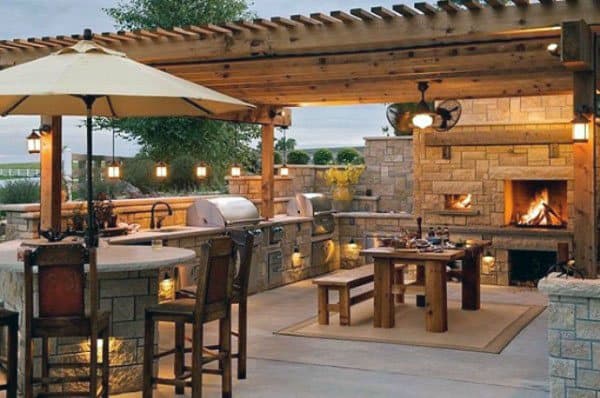 Stone pergola pavilion featuring an outdoor kitchen, bar seating, dining area, and a cozy fireplace.