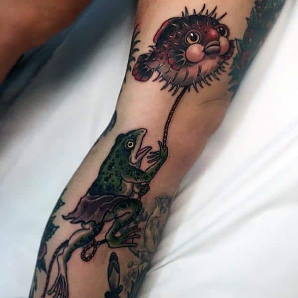 Whimsical pufferfish tattoo featuring a frog holding it like a balloon, blending fantasy and traditional tattoo elements with bold colors.
