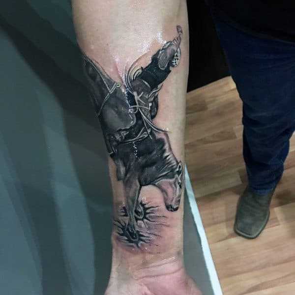 Black and grey tattoo of a bull rider in mid-air, detailed with intense shading and dynamic movement, placed on the forearm for impact.