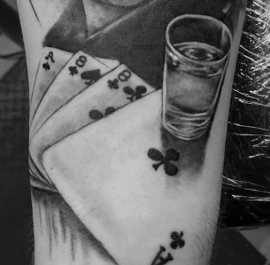 Black and grey shot glass tattoo featuring a realistic glass on top of playing cards, creating a bold and stylish gambling-inspired design.