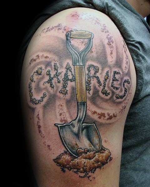 Realistic shovel tattoo with a wooden handle, digging into the ground, surrounded by dirt forming the name "Charles" in an earthy design.
