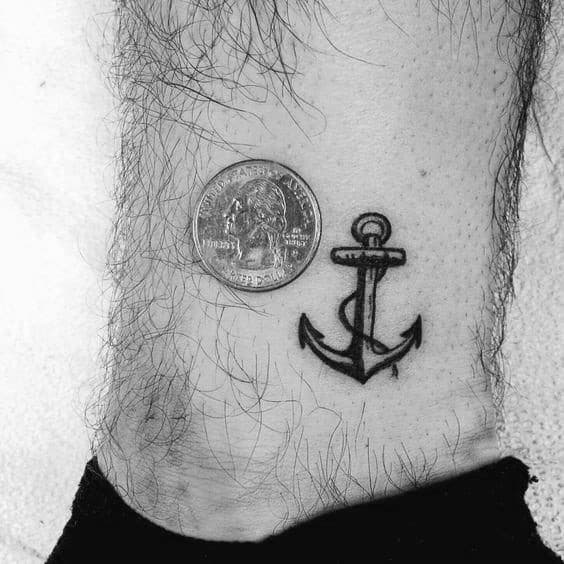 Tiny anchor tattoo on the ankle next to a quarter for scale, featuring fine black outlines and subtle shading, symbolizing stability and strength.